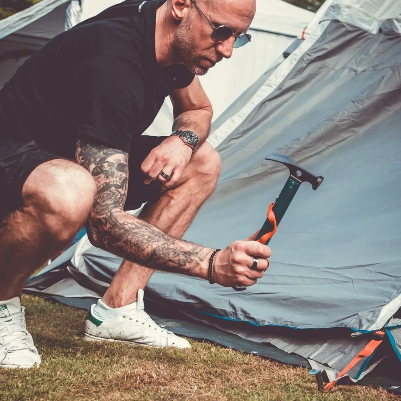 Ein Mann kniet auf Rasen und schlägt mit einem Hammer die Heringe eines Zeltes auf dem Campingplatz ein